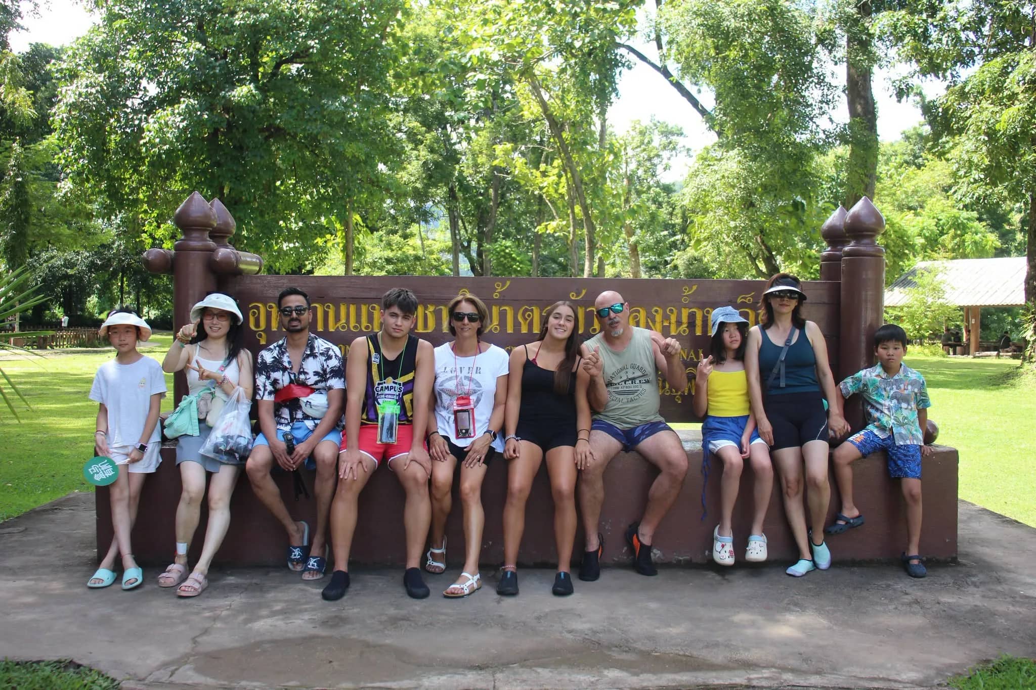 A group of visitors poses on a bench in front of the Bua Tong Sticky Waterfall park sign
