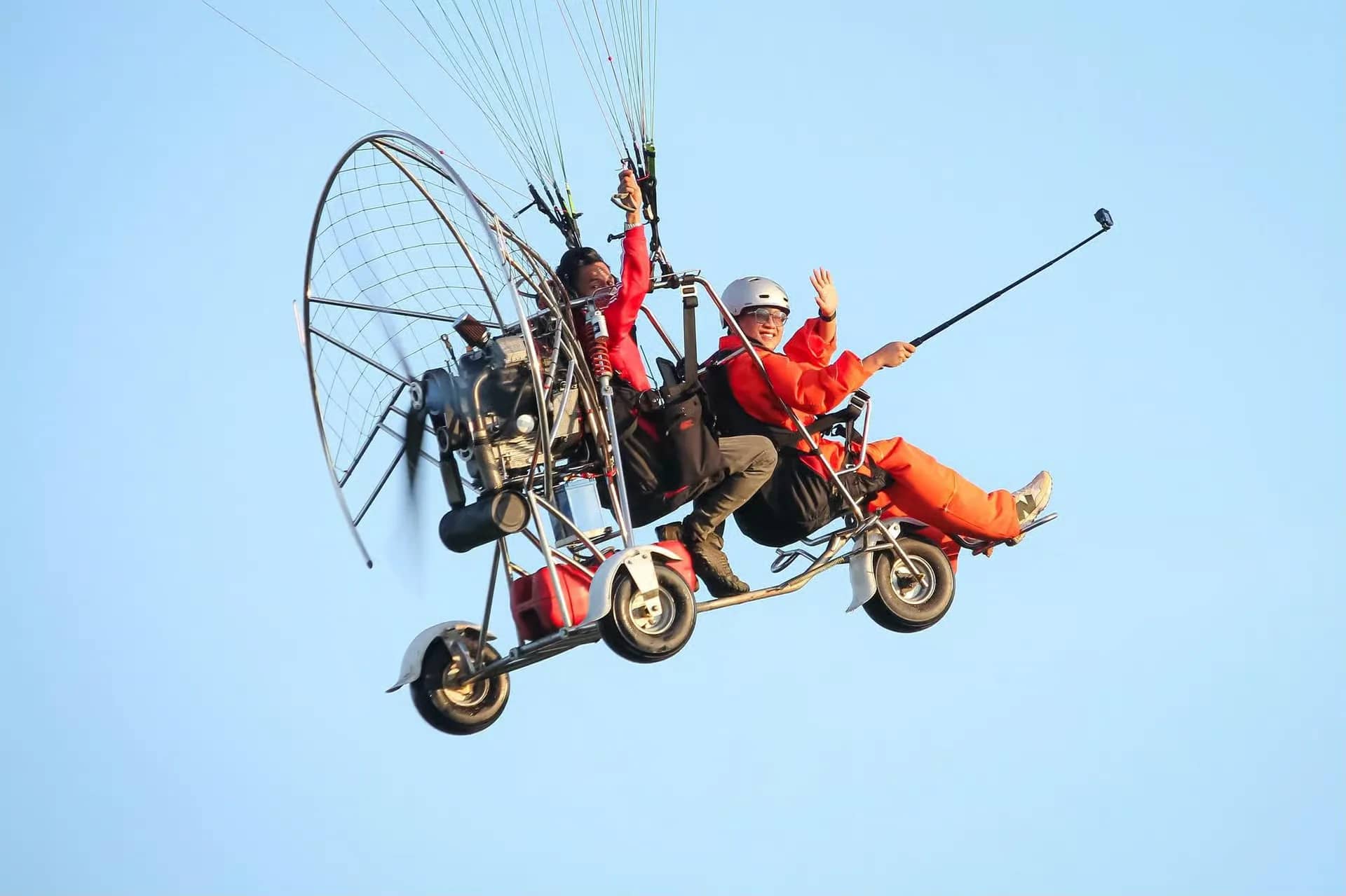 A passenger in an orange suit waves from a tandem paramotor trike while holding a selfie stick