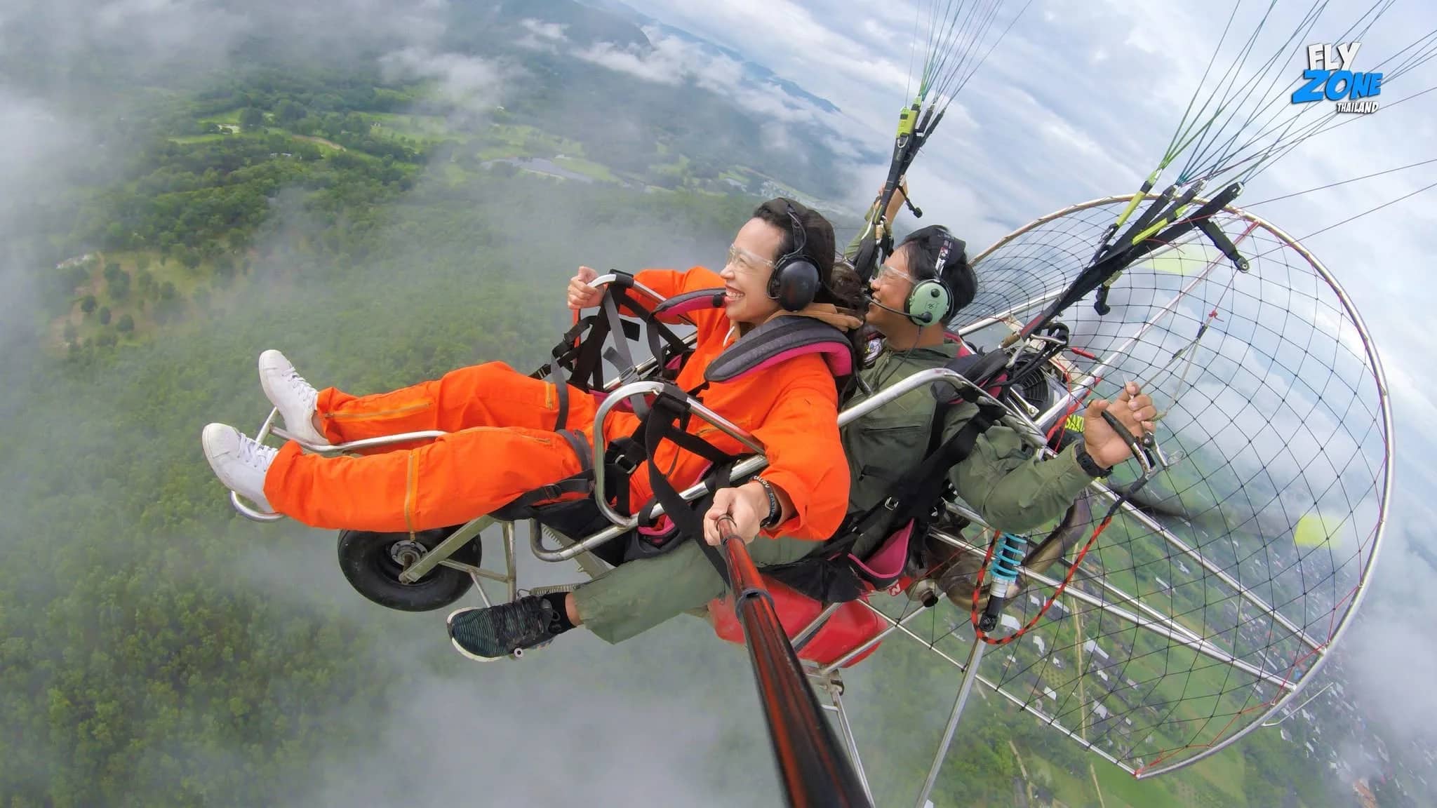 A pilot and passenger in an orange suit fly a paramotor high above clouds and green fields