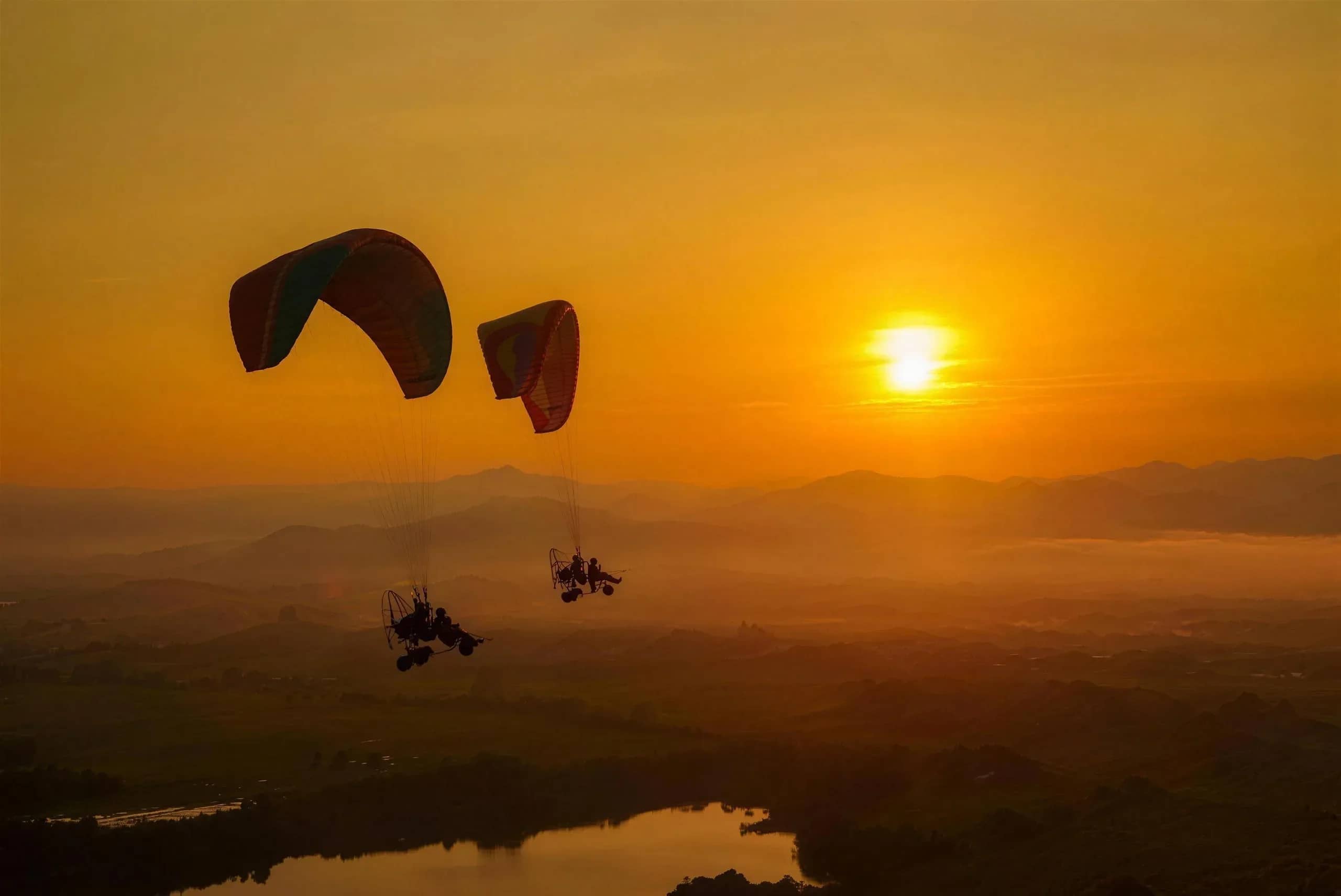 Two paramotors fly side by side across an orange sunset sky above a misty valley