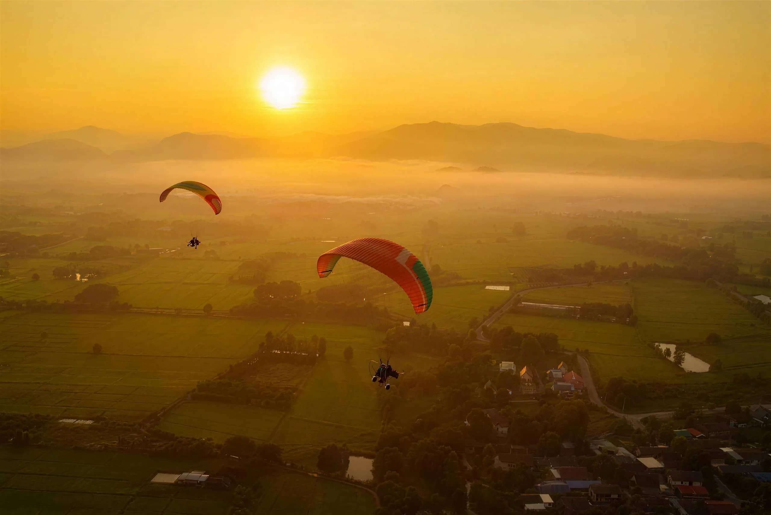 Paramotor wings drift above green rice fields and villages under a bright rising sun
