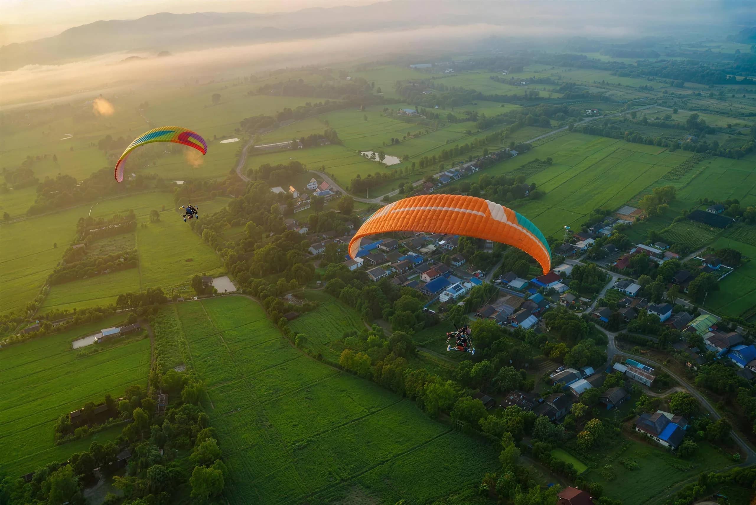 Two colourful paramotor wings fly over villages and green rice paddies on a misty morning
