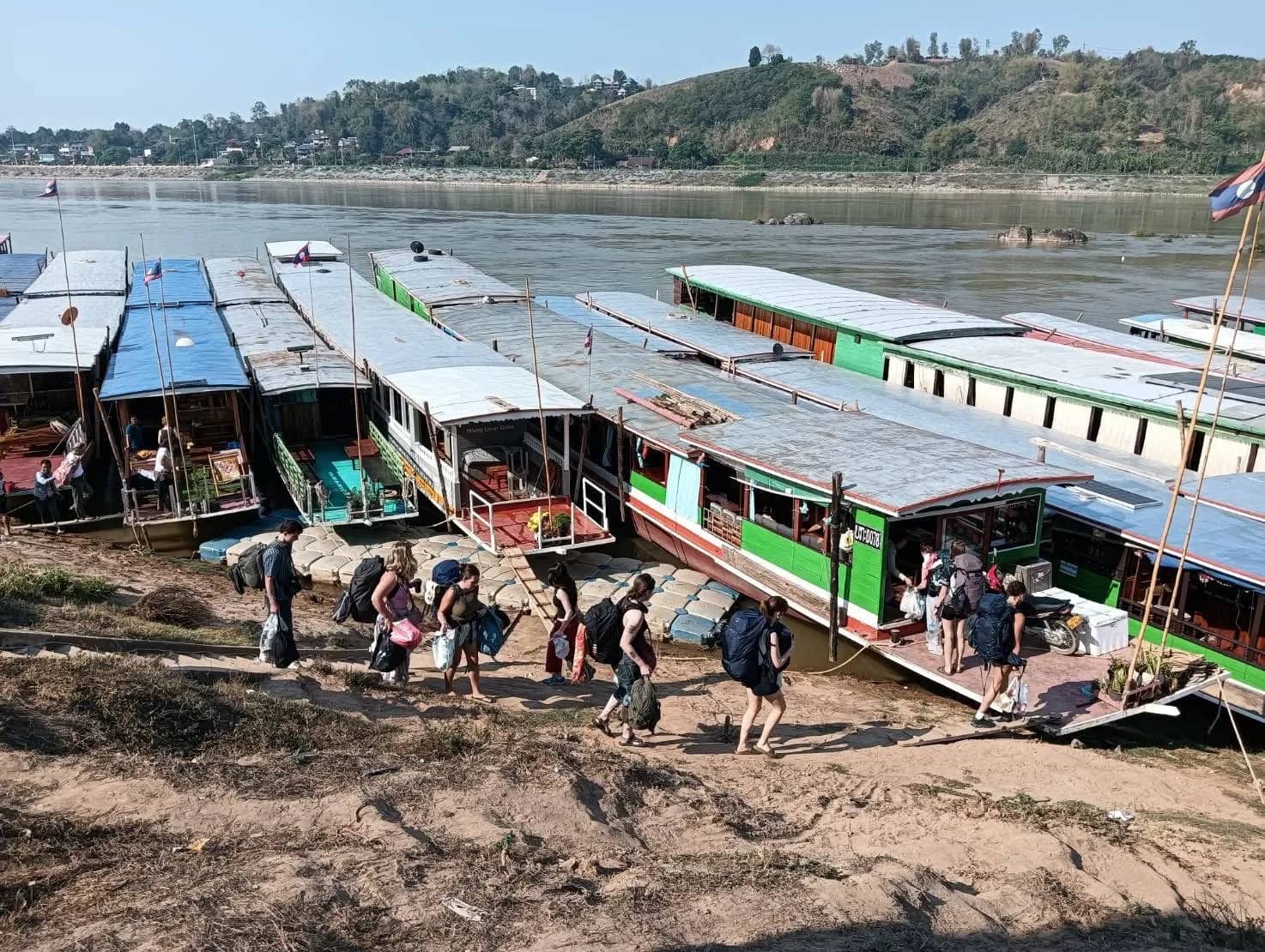 Travellers with backpacks walk down the riverbank to a row of moored slow boats on the Mekong