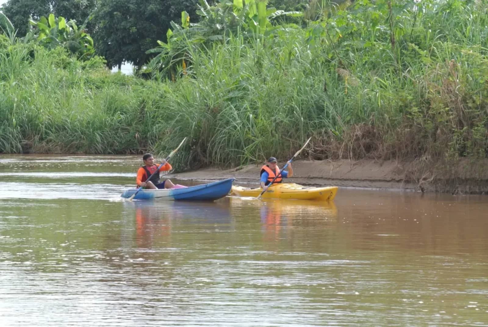 Two paddlers in life jackets kayak along a calm river bordered by tall green grasses