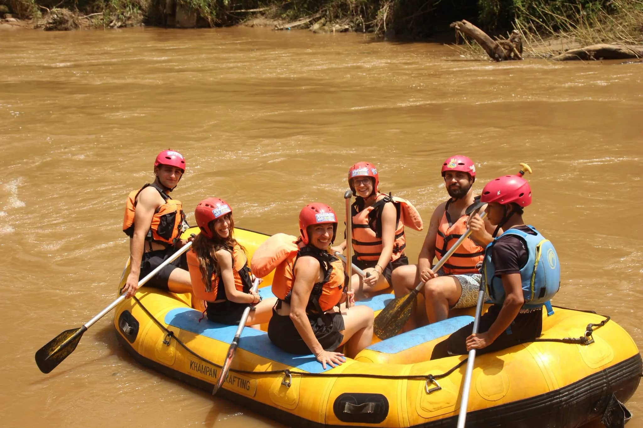 Smiling rafters in red helmets and life jackets sit in a yellow raft on a brown river
