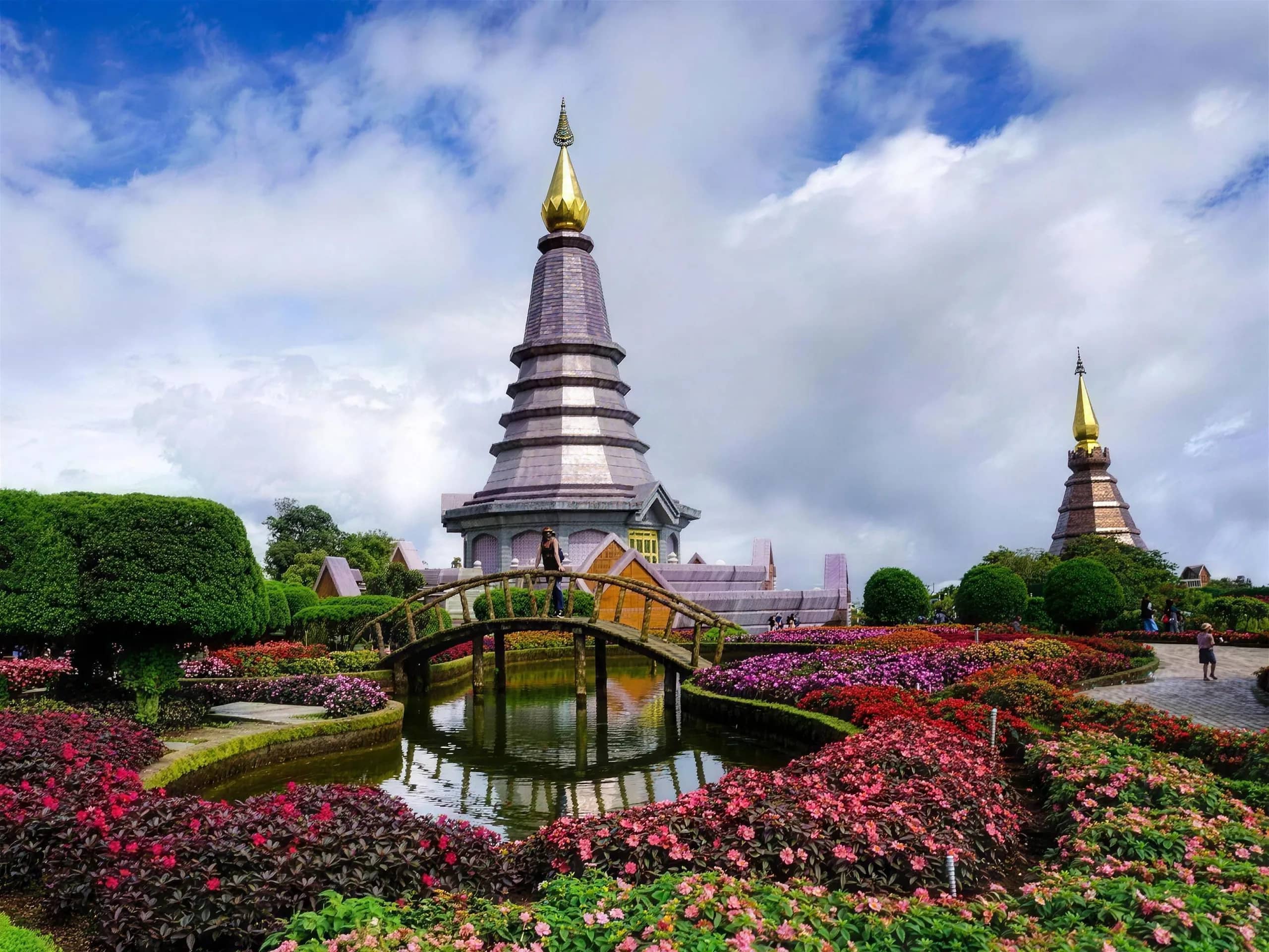 Pink and red flower beds and a small bridge lead to the royal pagodas under a cloudy sky.