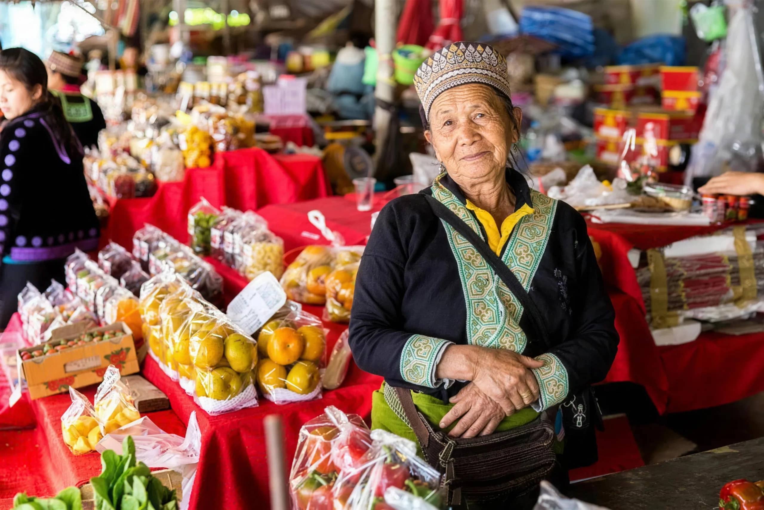 A vendor in an embroidered jacket and hat stands beside a stall of fruit and snacks