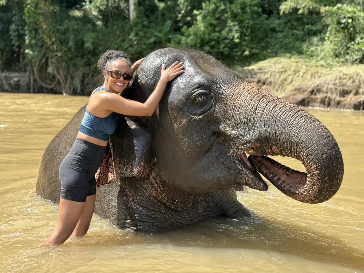 A visitor in swimwear hugs the head of an elephant lying in a shallow river at Pon Elephant Sanctuary