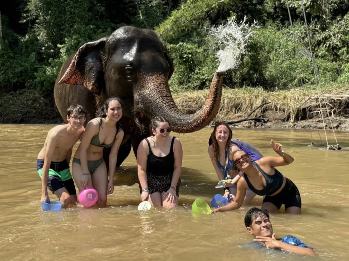 An elephant sprays water from its trunk as guests pose in the river at Pon Elephant Sanctuary