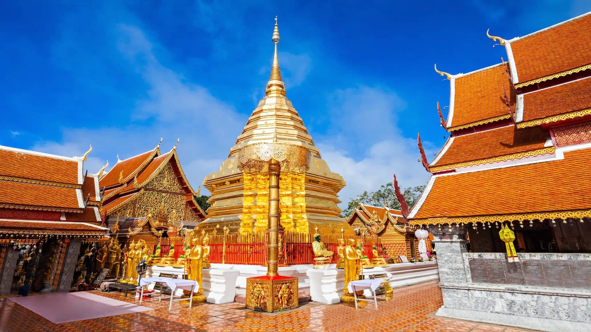The golden chedi of Wat Phra That Doi Suthep stands between red-roofed temple halls under blue sky.
