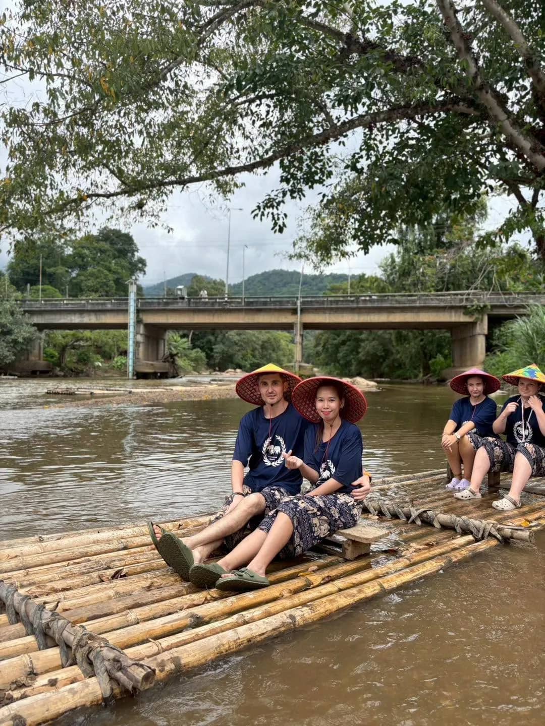 Guests in straw hats sit on a bamboo raft on a brown river below a road bridge