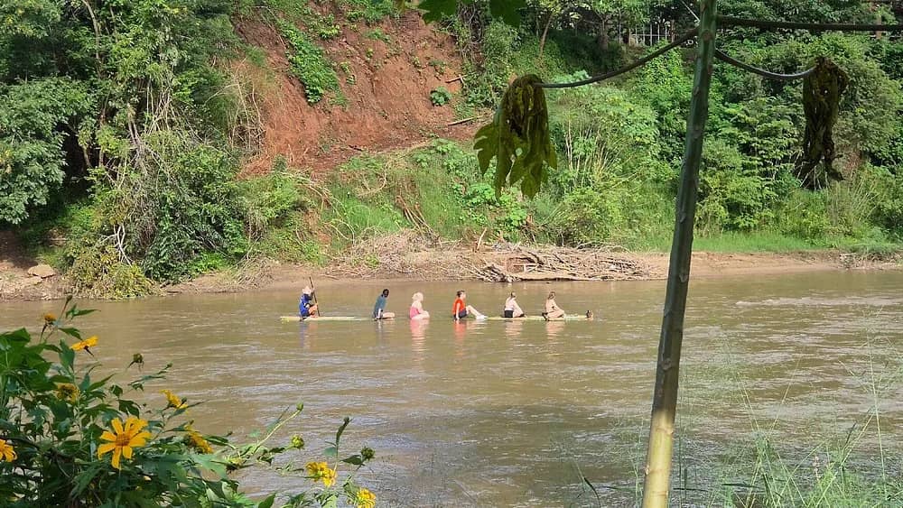A line of guests drifts down a broad brown river on a long bamboo raft below green hills.