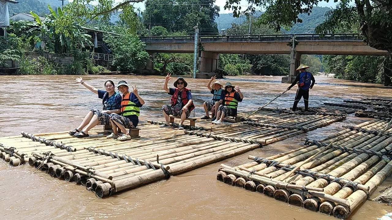 Guests in life jackets wave from bamboo rafts moored under a bridge while a guide holds a pole.