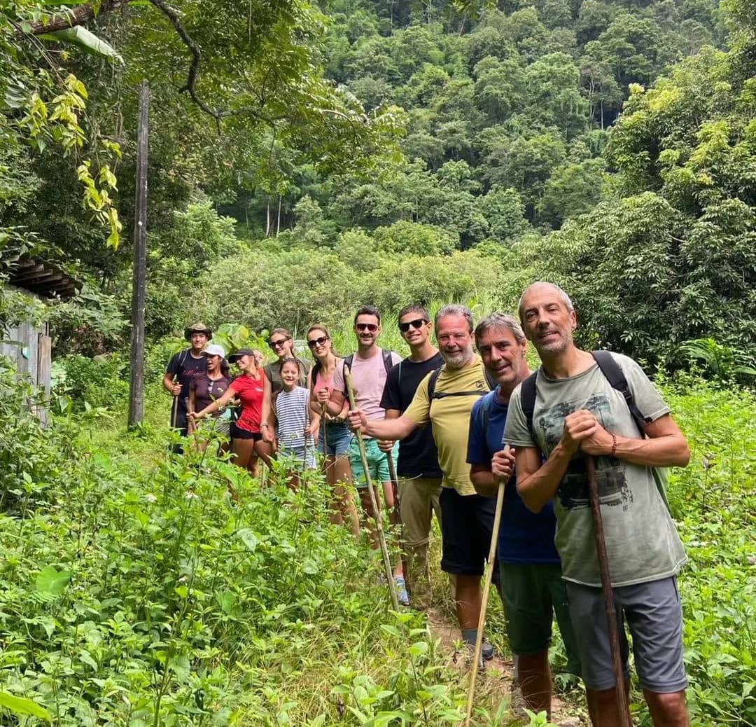 A group of hikers with walking sticks smiles on a narrow path through thick green jungle