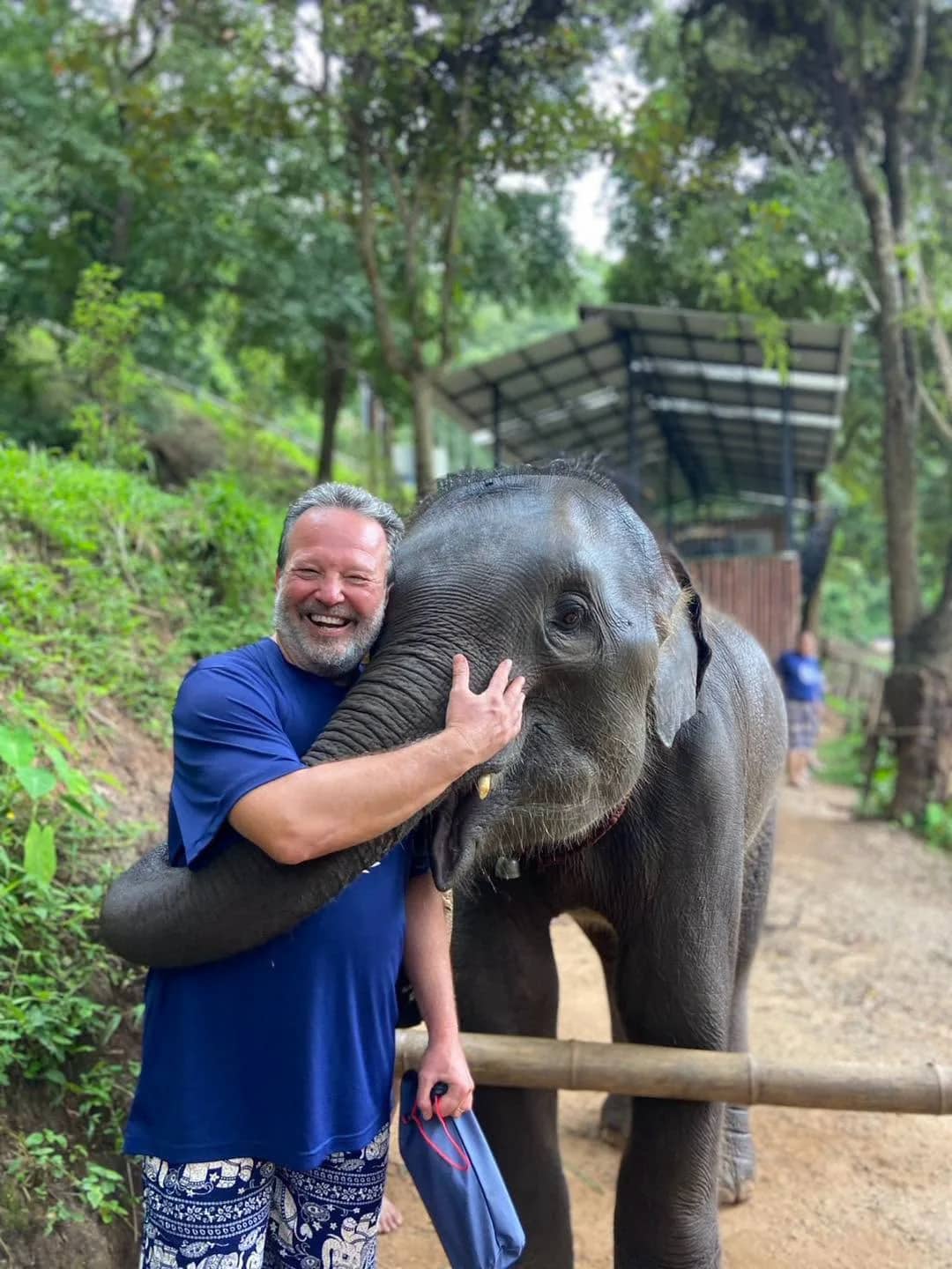 A smiling visitor gets a hug from a baby elephant on a forest path