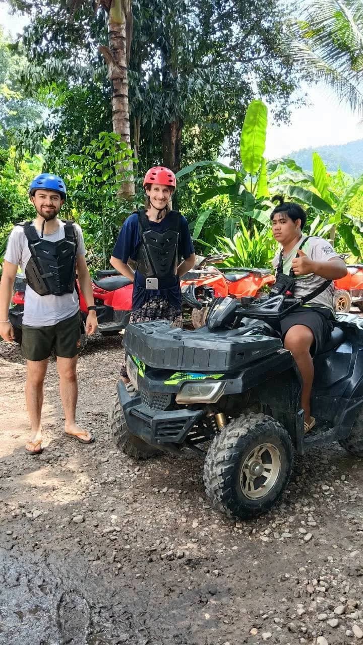 Three riders in helmets and body armour pose beside a black ATV on a muddy track