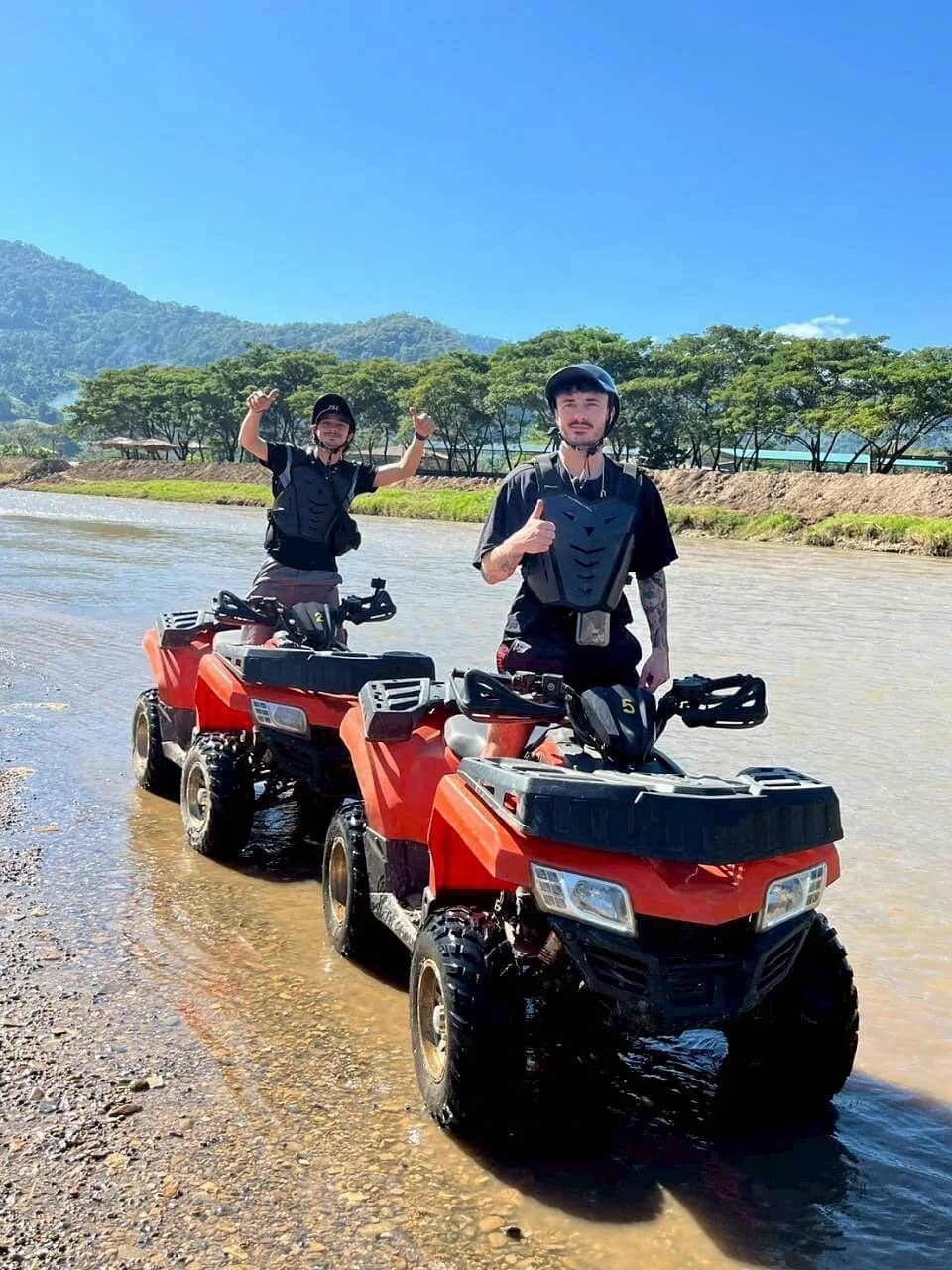 Two riders stand on red ATVs parked in a shallow river with hills behind