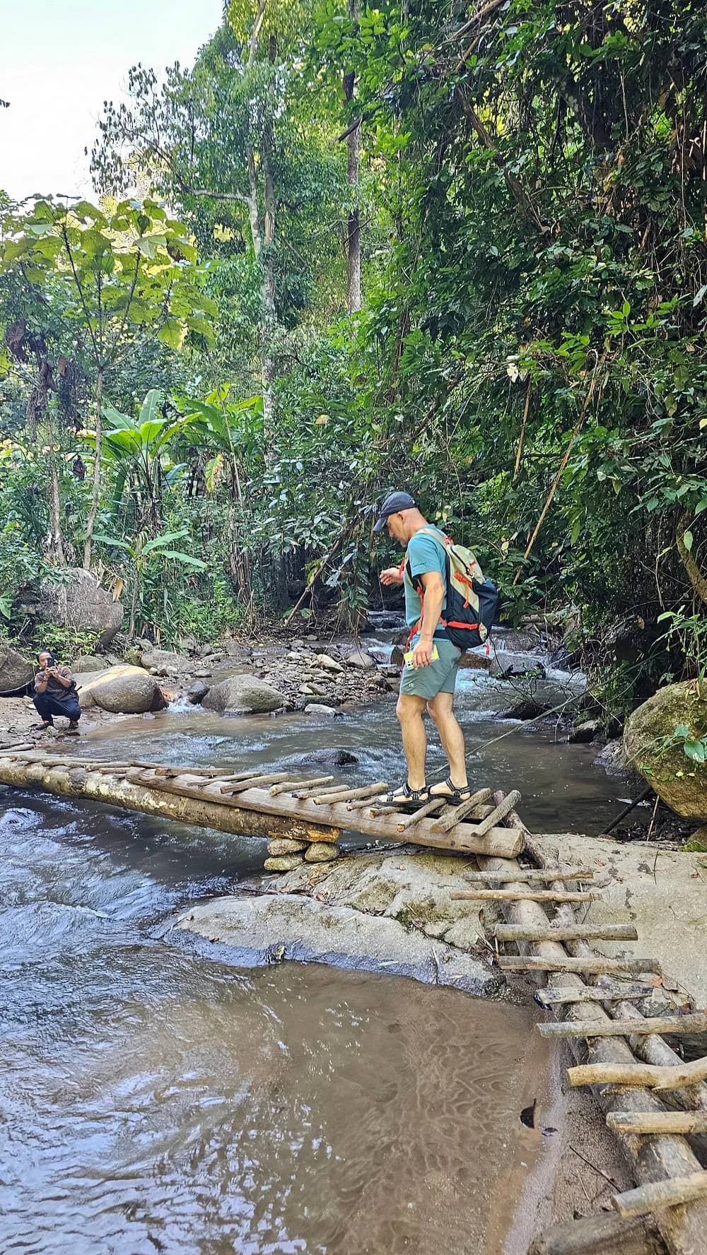 A hiker crosses a bamboo bridge over a rocky jungle stream