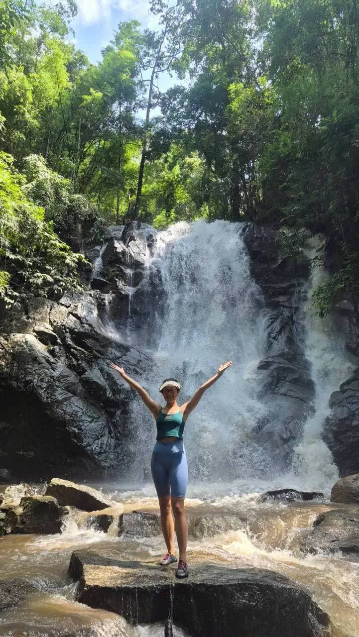 A hiker raises both arms on a rock in front of a tall waterfall in the jungle