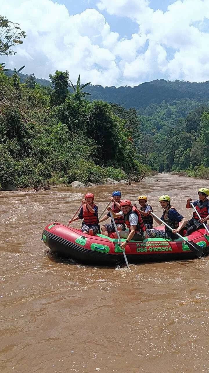 A red raft full of helmeted rafters paddles down a muddy river in a green mountain valley