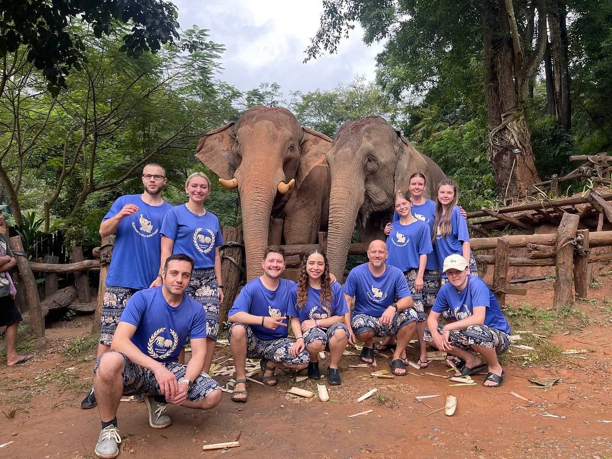 A group of visitors in blue shirts poses in front of two elephants at Elephant In Wild Sanctuary