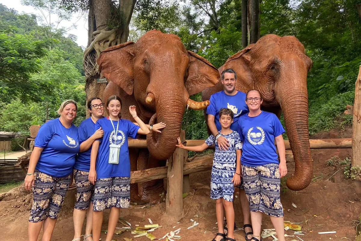 Visitors in matching blue shirts pose with two elephants covered in red dust at the sanctuary.
