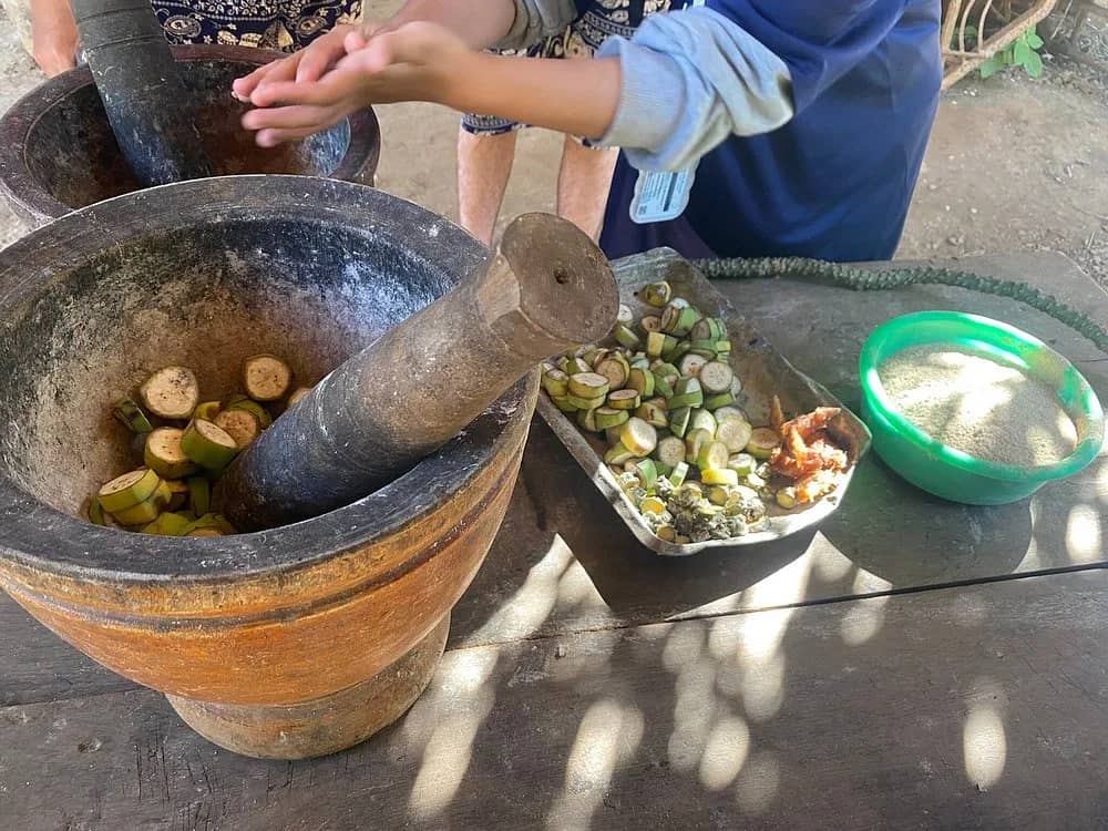 A stone mortar and pestle with sliced bananas beside a tray of chopped ingredients on a table.