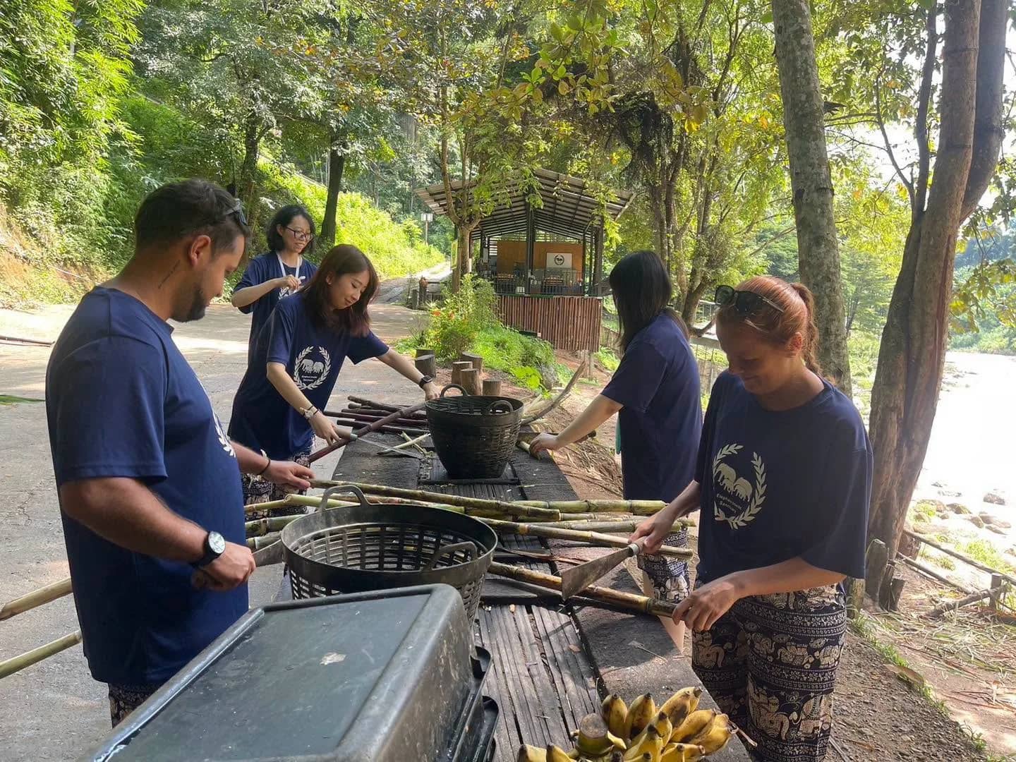 Visitors in blue shirts cut sugarcane and bamboo at a wooden table beside a river.