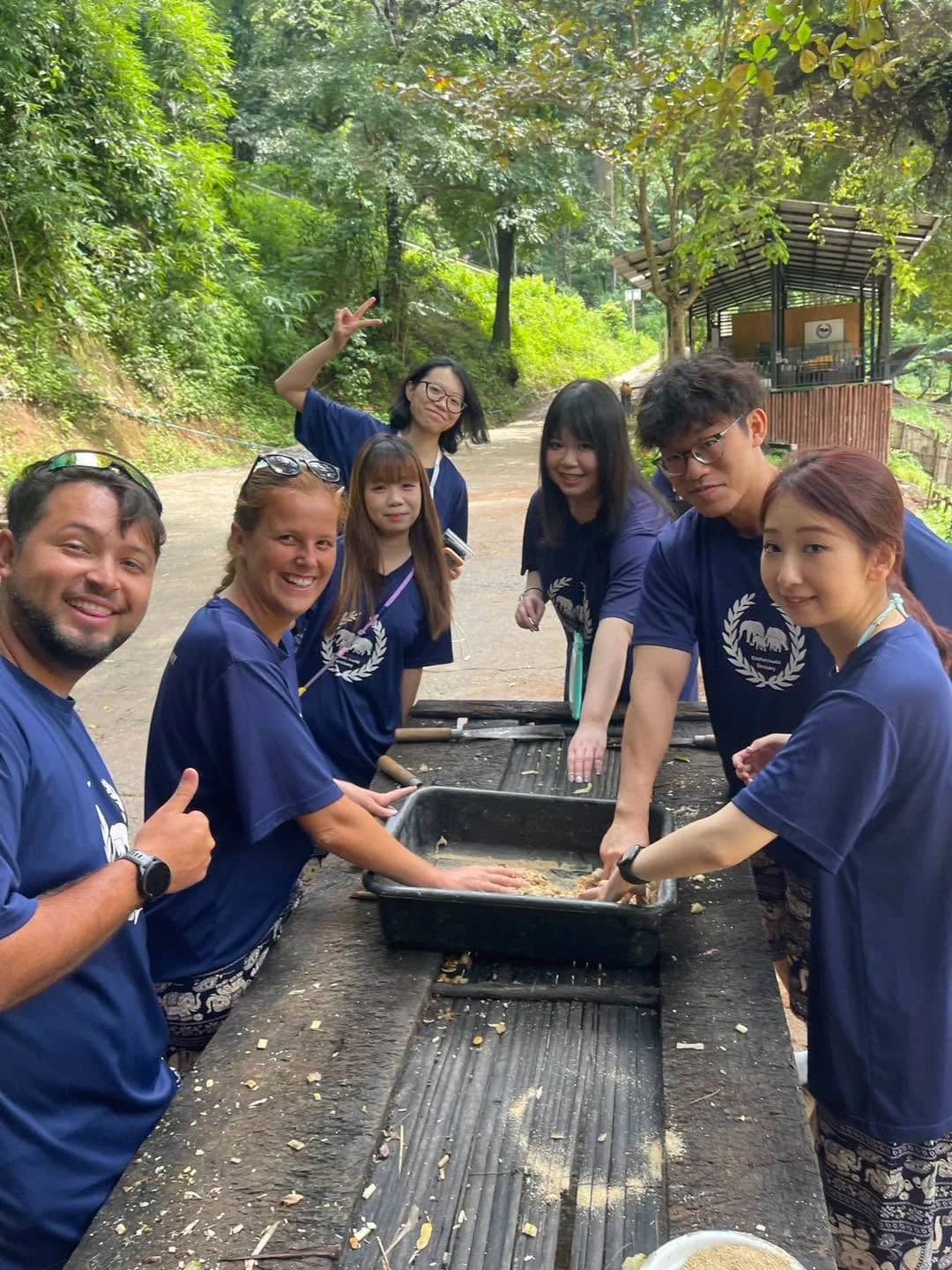 Smiling visitors in blue shirts mix food in a metal tray at a long wooden trough.