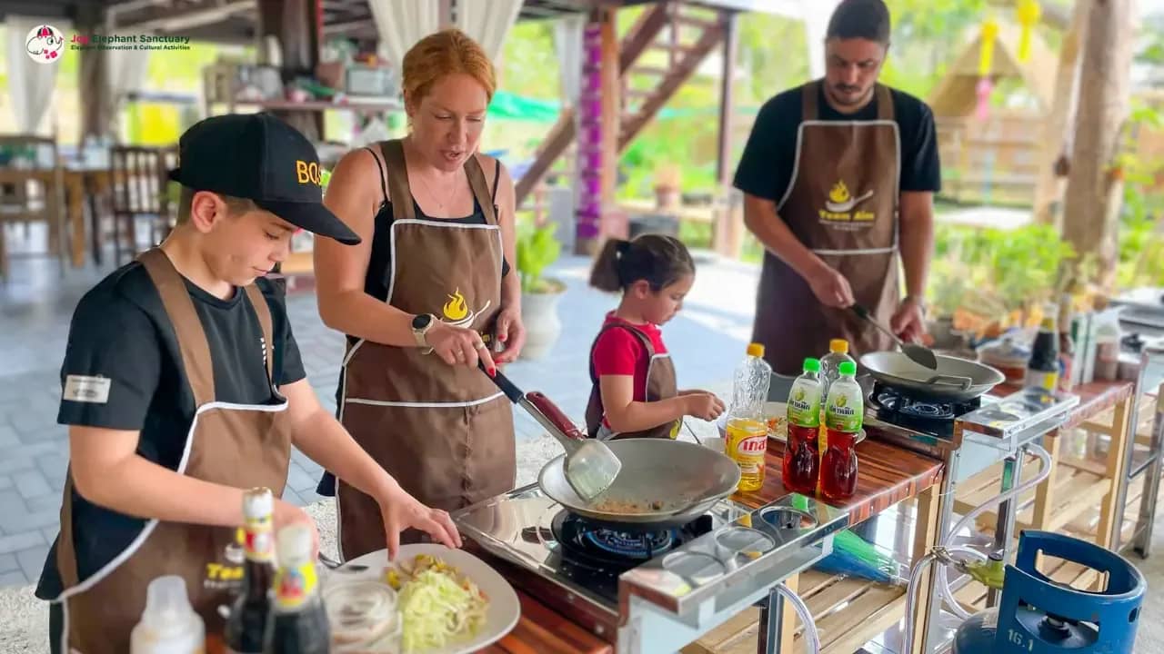 A family in brown aprons cooks with woks at outdoor stoves in an open-air kitchen