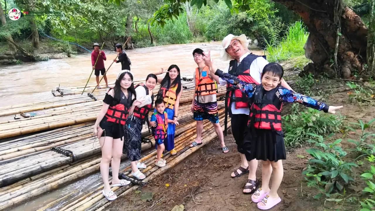 A family in life jackets poses on bamboo rafts lined up at a riverbank before rafting