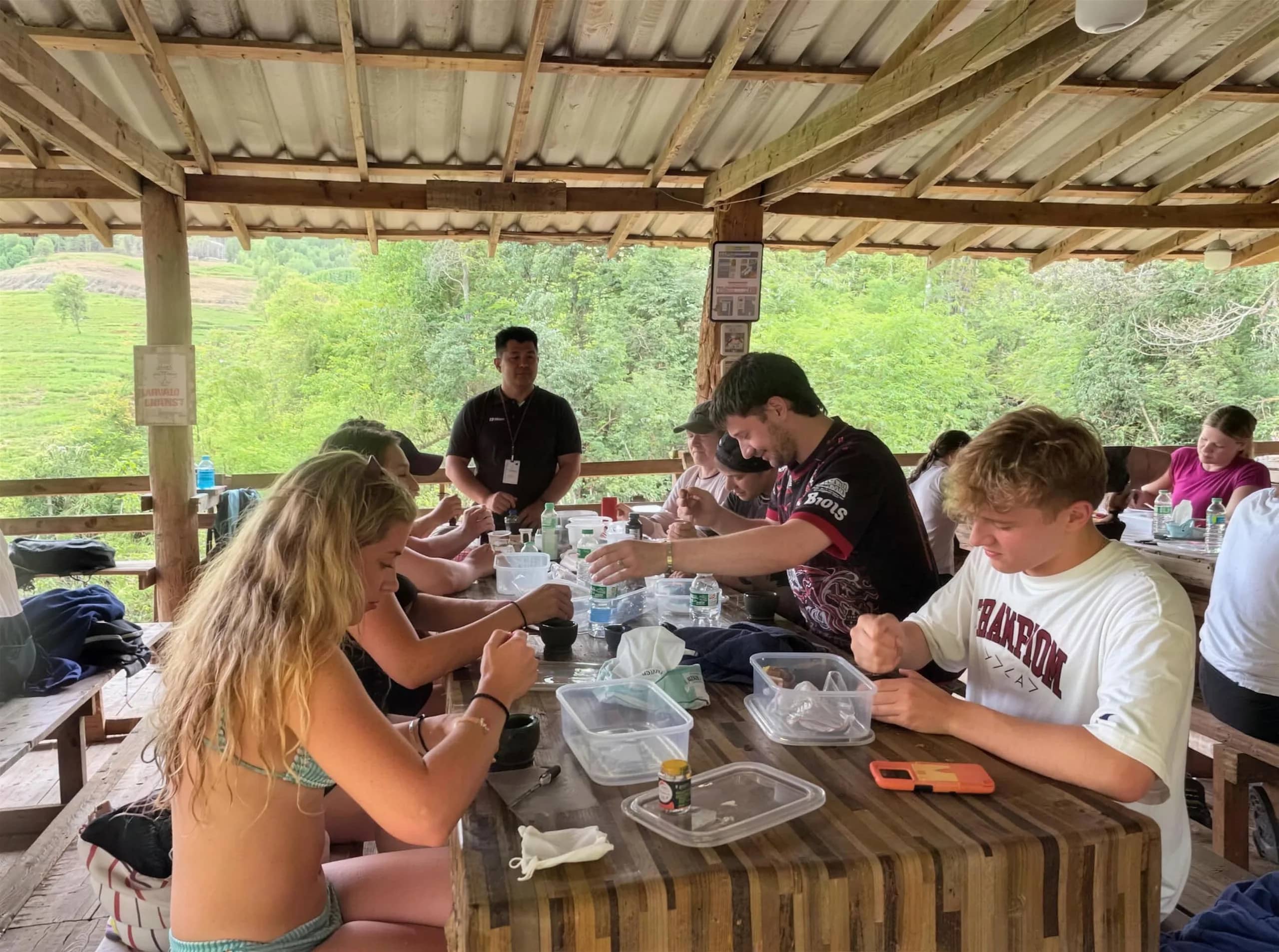 Visitors work with small bowls and containers at a wooden table under a tin-roofed shelter in the forest