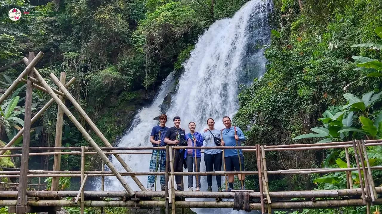 Four hikers stand on a bamboo bridge in front of a tall jungle waterfall near Doi Inthanon.