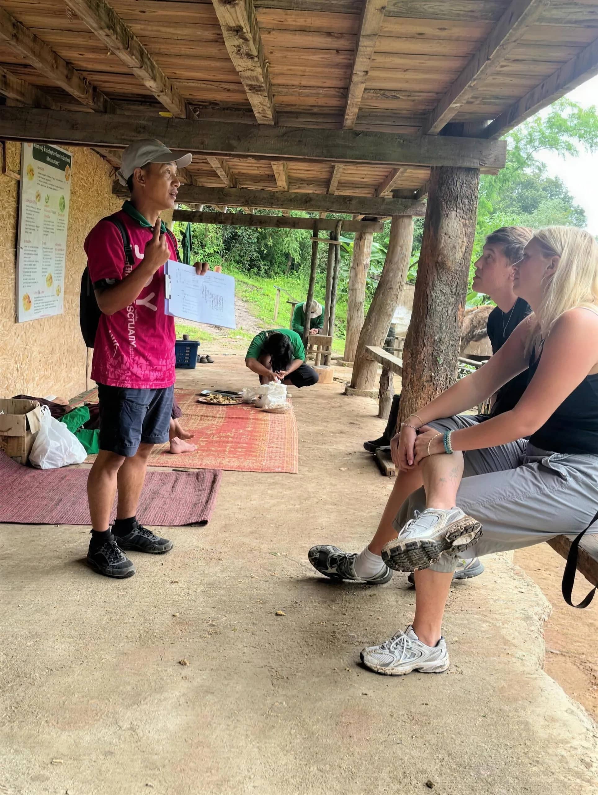 A guide in a pink shirt talks to seated visitors under a wooden shelter at the elephant camp