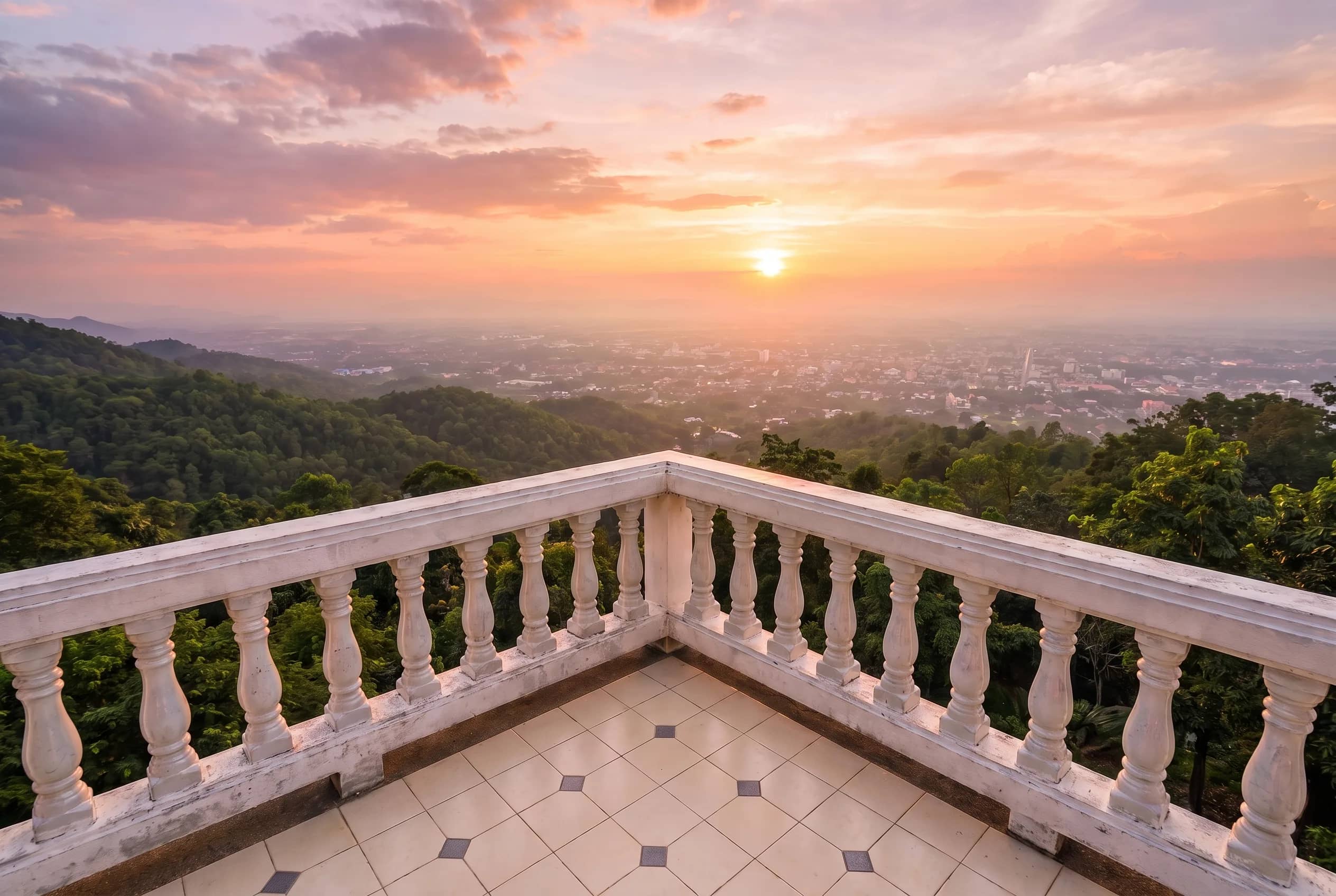 A white balustrade terrace overlooks forested hills and the city of Chiang Mai at sunset