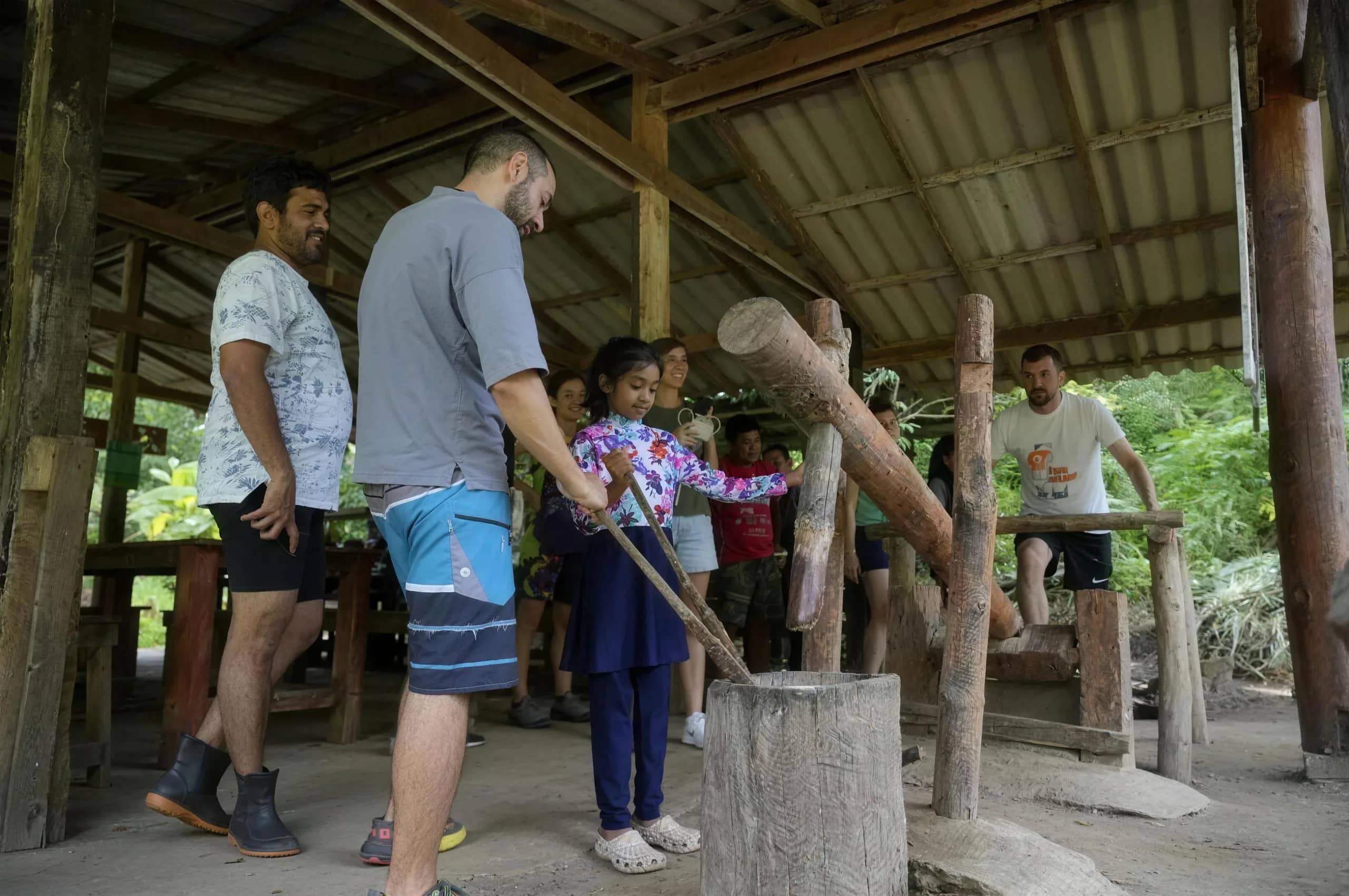 Visitors take turns pounding with a large wooden pestle in an open-air sala at the sanctuary