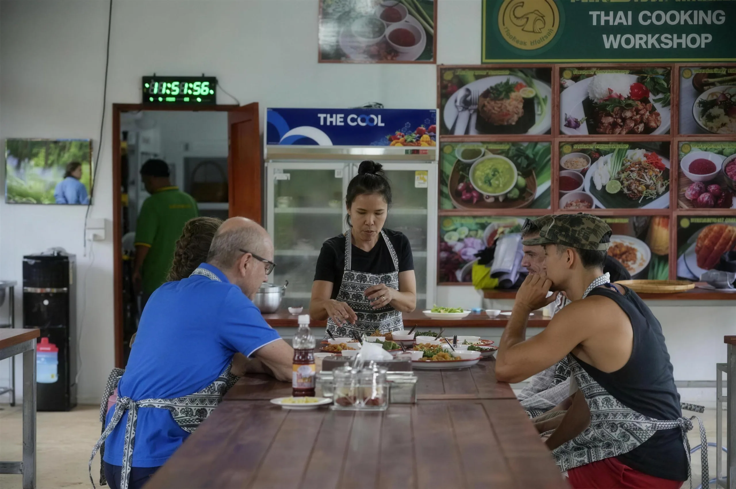 Guests in aprons sit at a long wooden table with dishes at the Thai cooking workshop