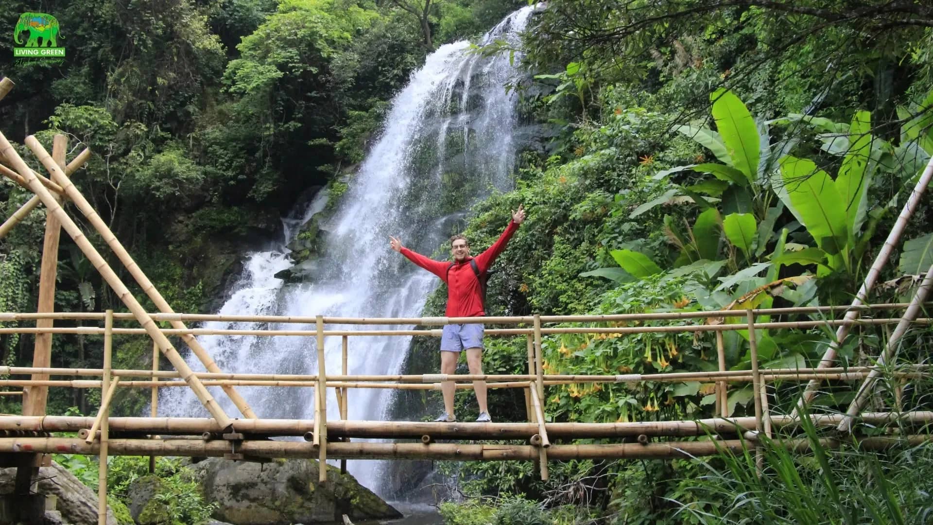 A hiker stands with arms raised on a bamboo bridge in front of a forest waterfall at Doi Inthanon