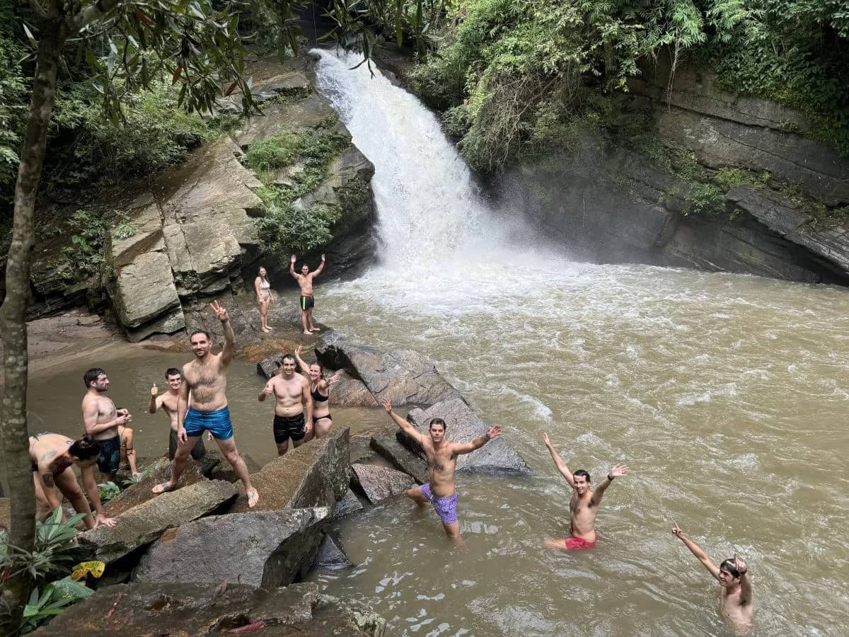 A cheerful group swims and stands on rocks in the pool below a rushing waterfall