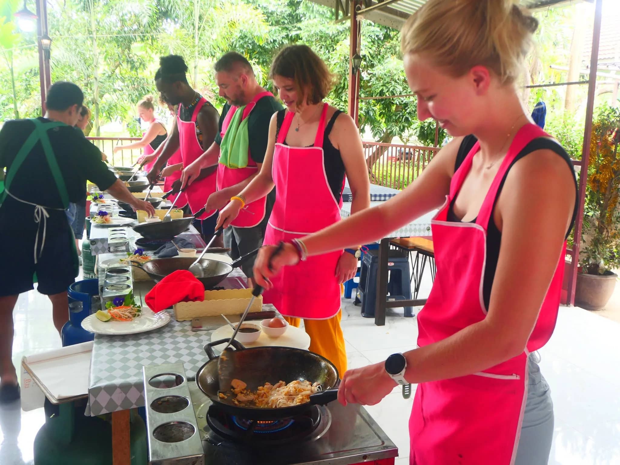 Guests in pink aprons stir-fry dishes in woks at individual gas stoves in an open-air kitchen.