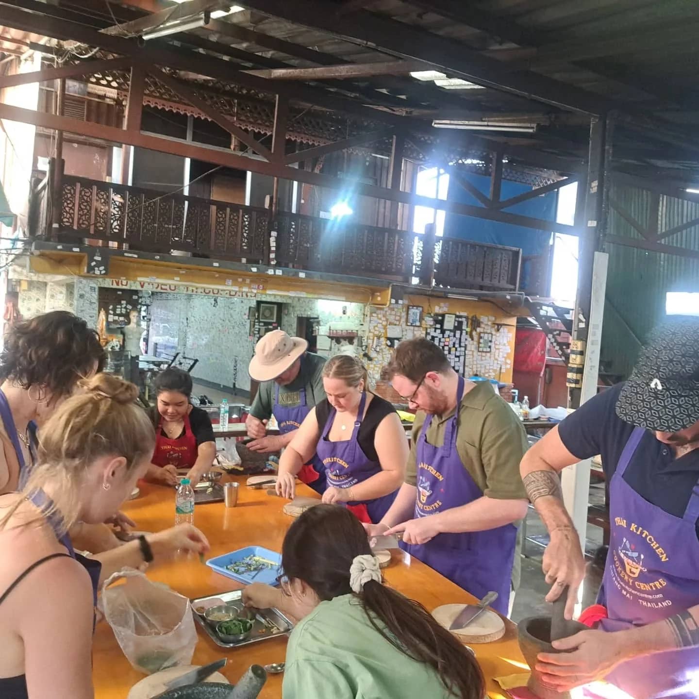 Students in aprons prepare ingredients together at a long table in a busy cooking school kitchen