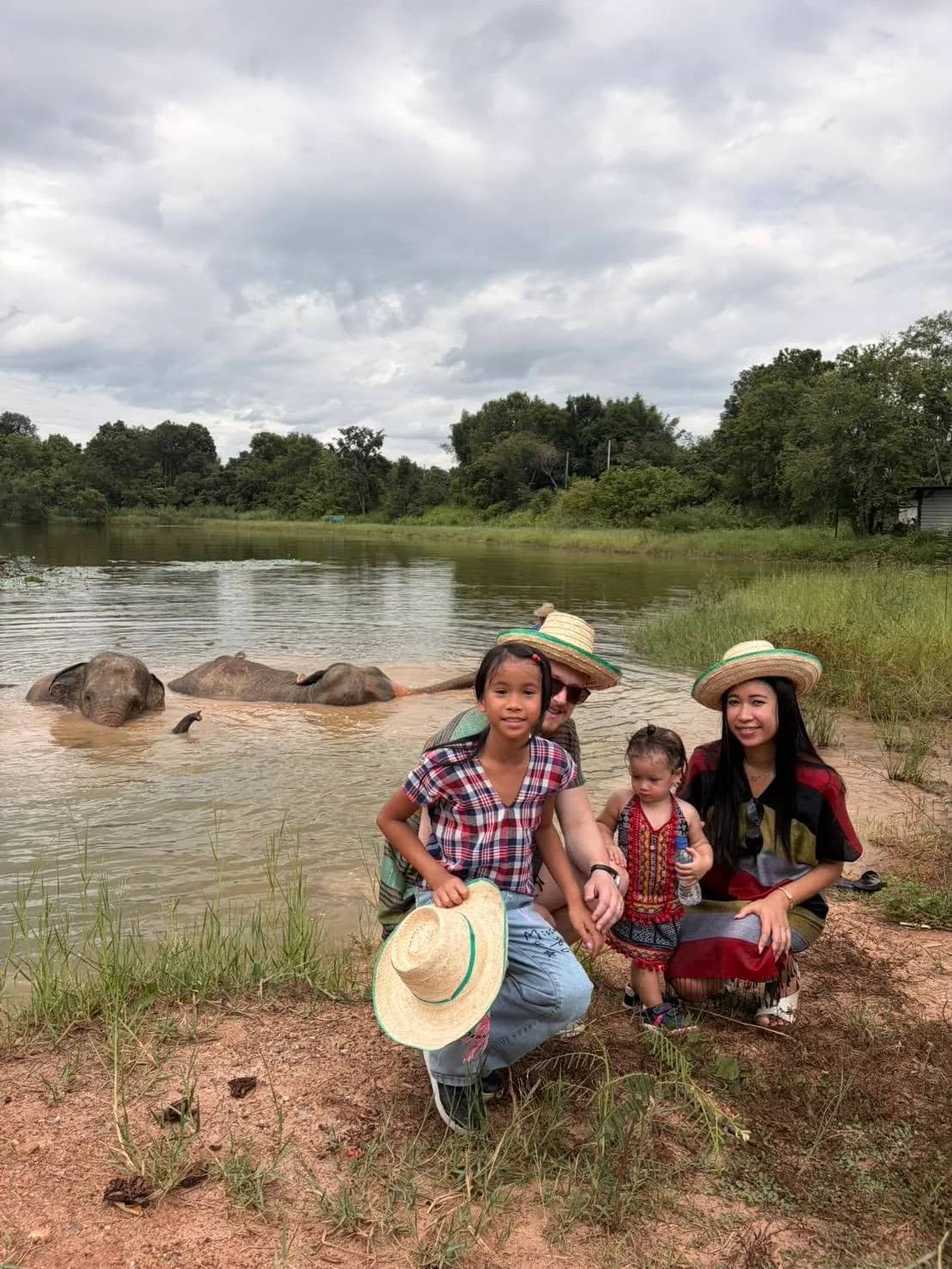 A family in straw hats poses on the bank while elephants bathe in the lake behind them