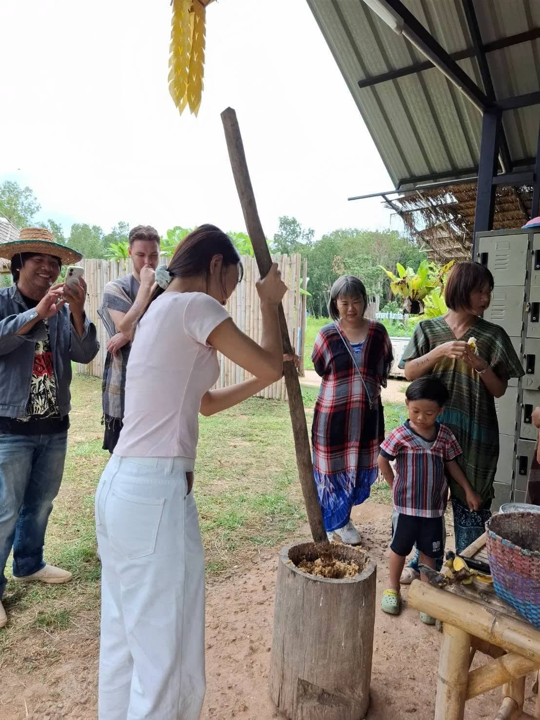 A visitor pounds a large wooden pestle into a mortar as other guests watch and take photos