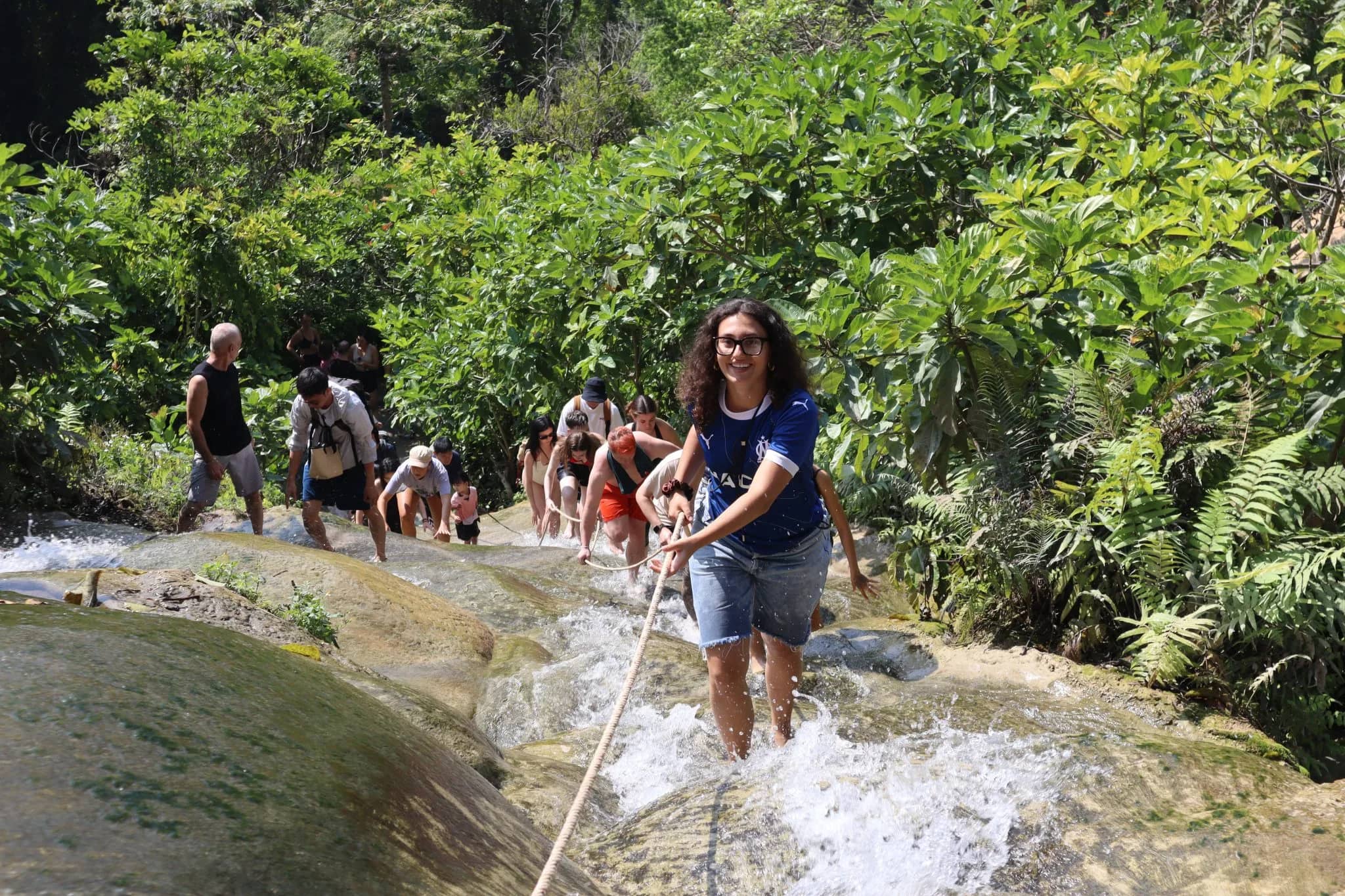 Visitors climb up the Sticky Waterfall holding a rope as water flows over the rocks