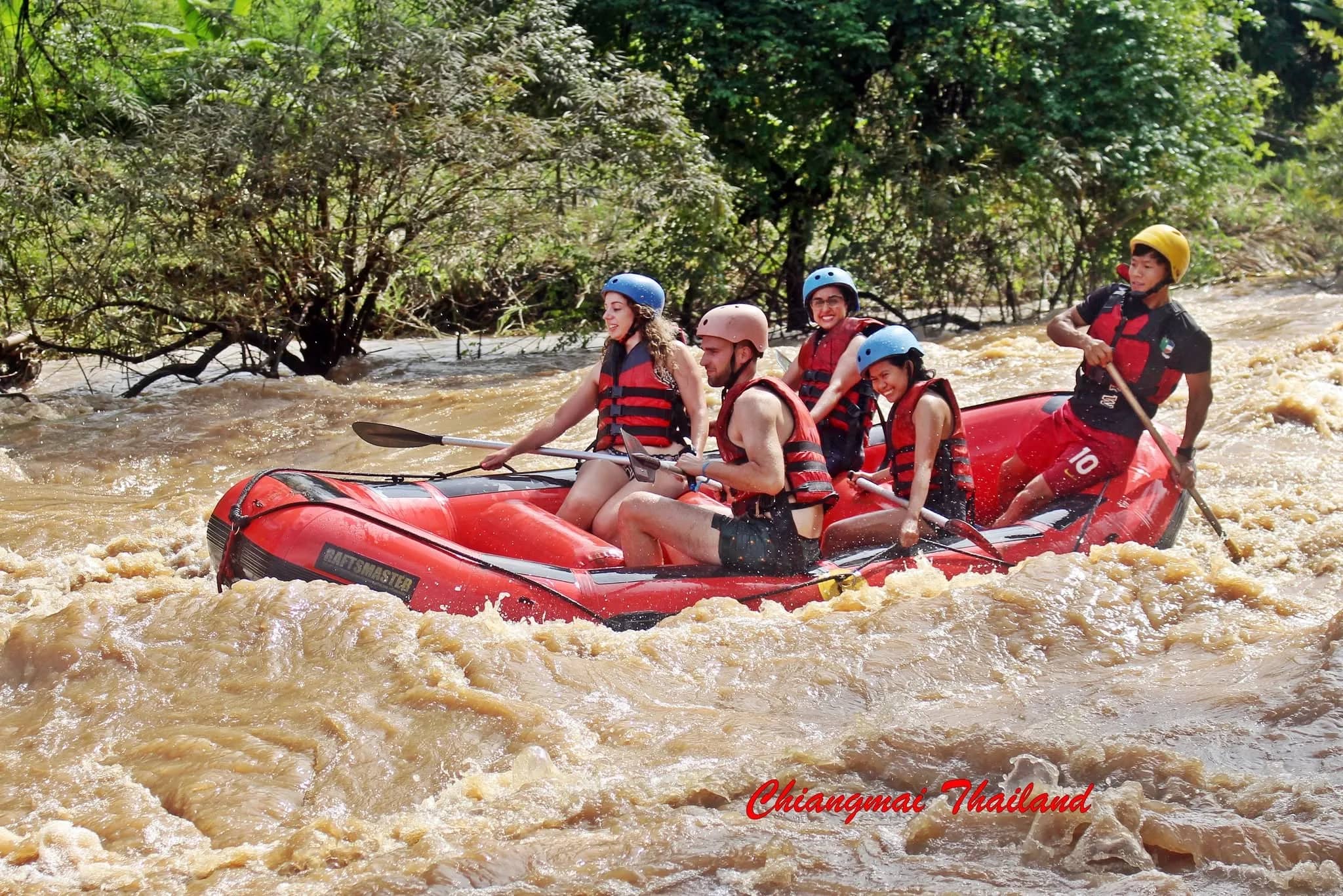 Guests in helmets and life jackets paddle a red raft through brown rapids as a guide steers at the back.