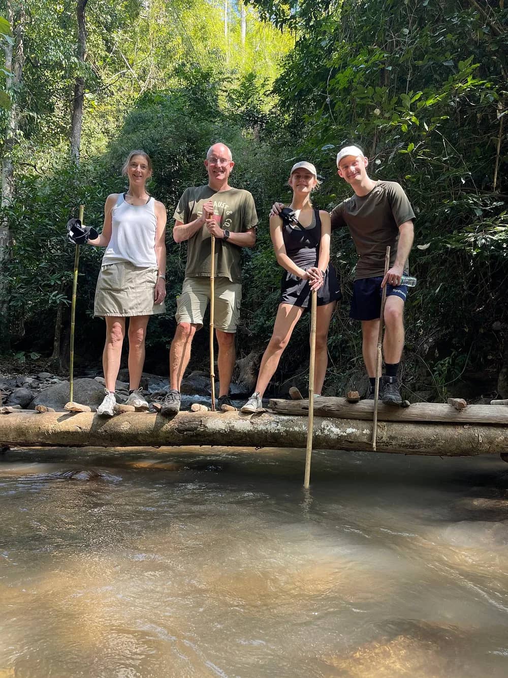 Four hikers holding bamboo walking sticks stand on a log bridge over a clear jungle stream.