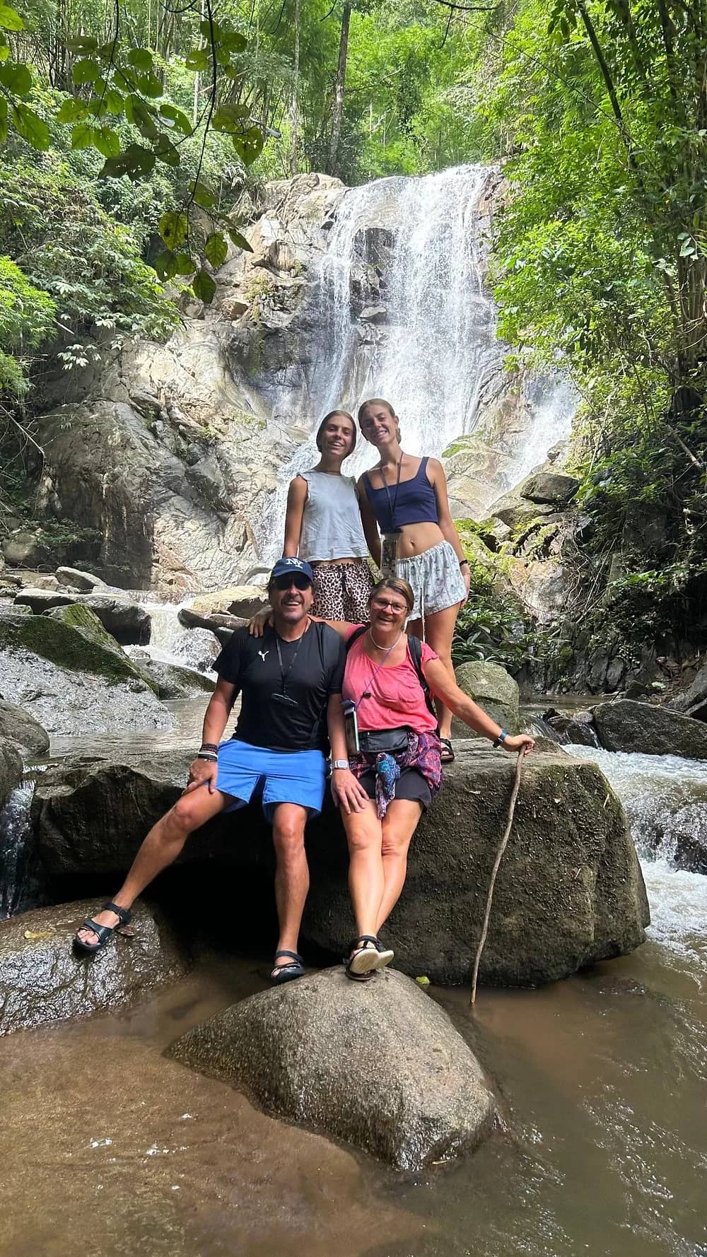 Four visitors pose on rocks at the foot of a forest waterfall during the trek.