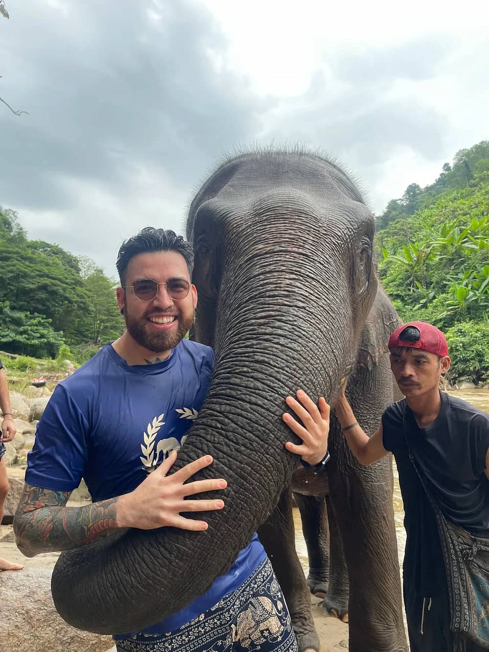 A smiling visitor wraps his arms around an elephant's trunk as a mahout stands beside them.