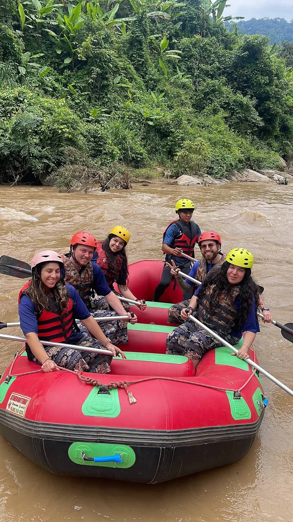 Smiling rafters in helmets and life jackets sit in a red inflatable raft on a jungle river.