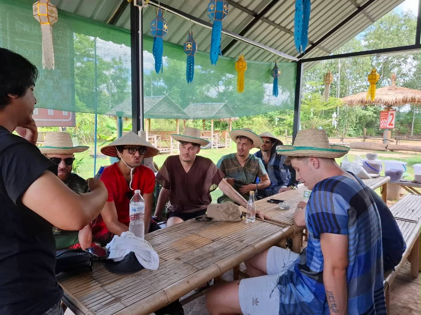 A guide talks to guests in straw hats seated at a bamboo table under a shelter